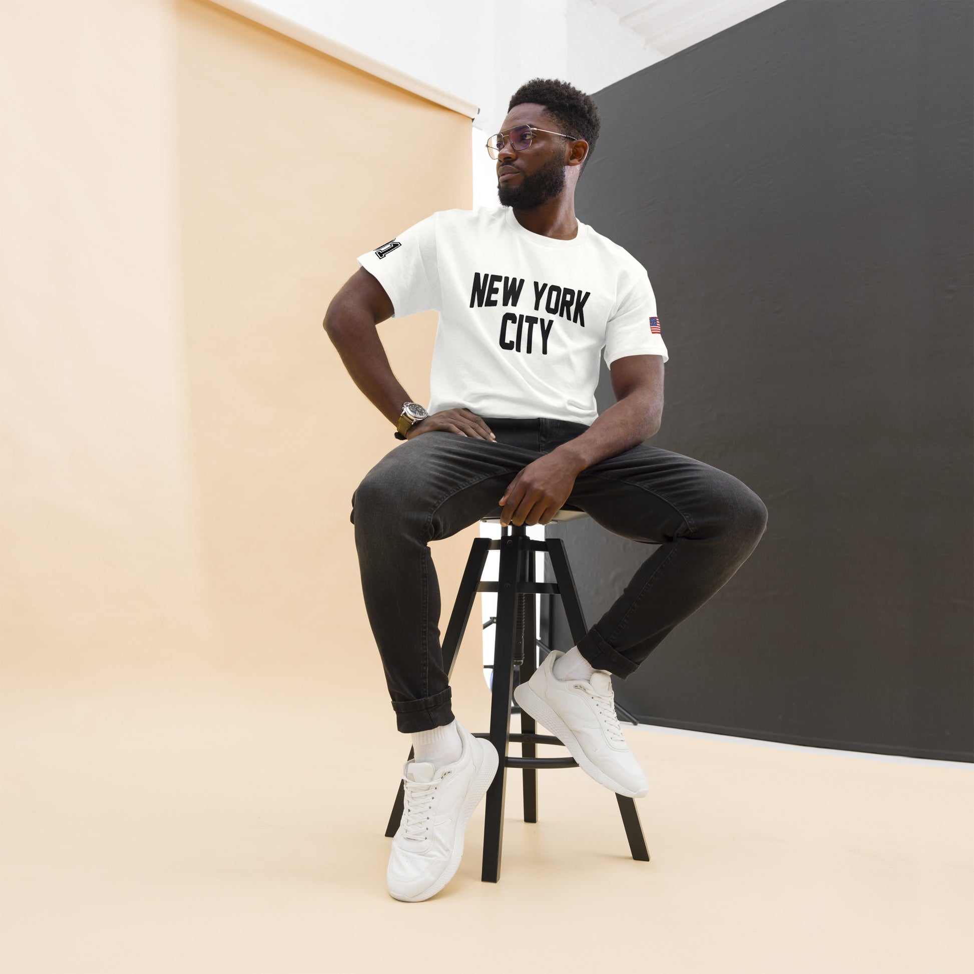 Man sitting on a stool wearing a 'New York City' t-shirt against a plain background