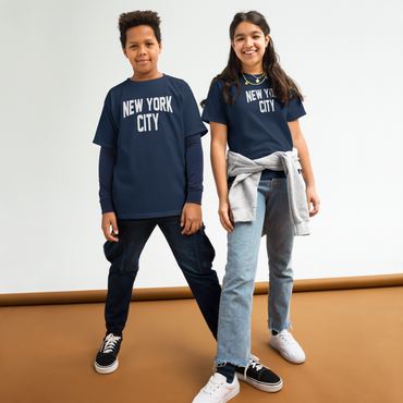 Two children wearing 'New York City' t-shirts standing on a brown floor with a white wall background.