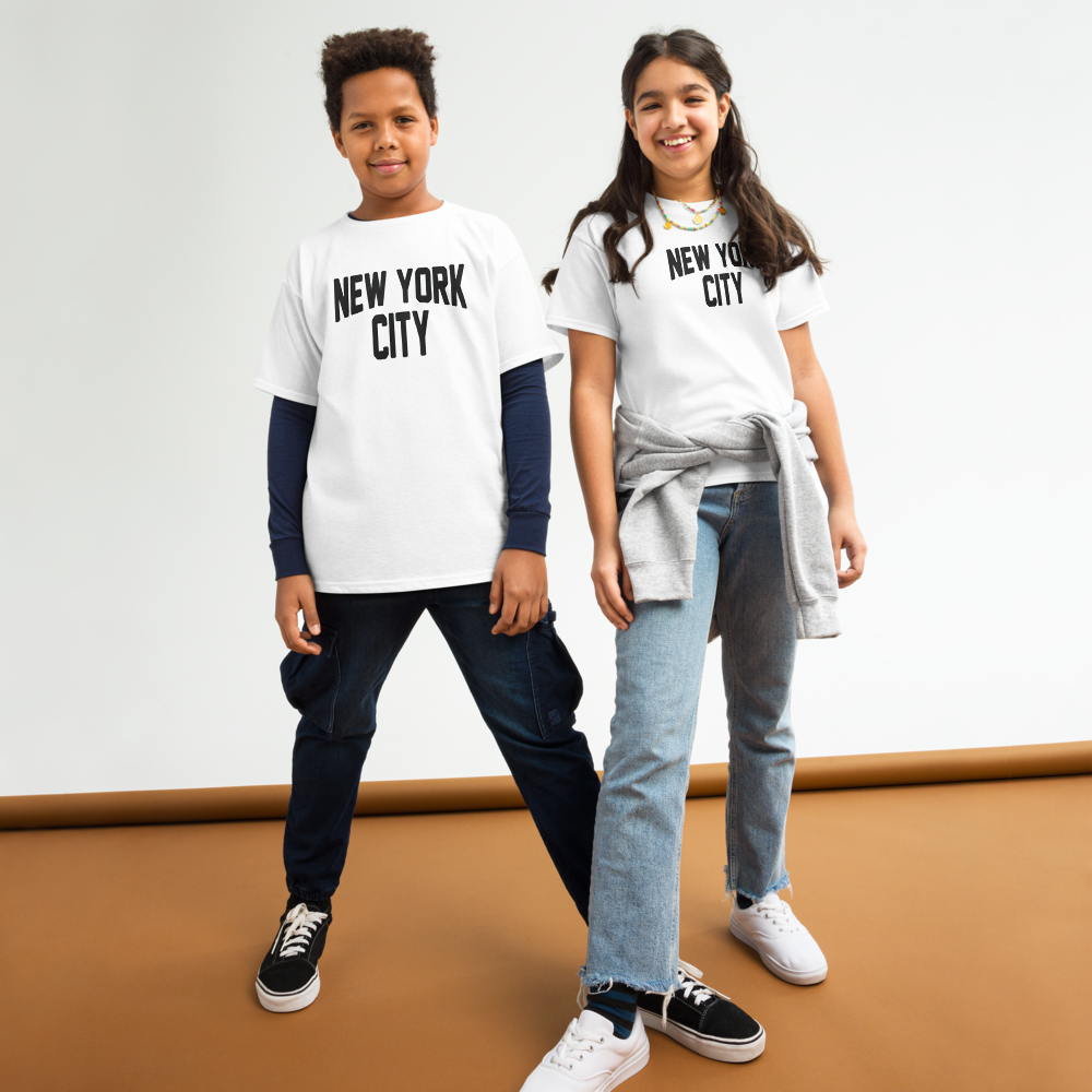 Two children wearing 'New York City' t-shirts standing on a brown floor with a white wall background.