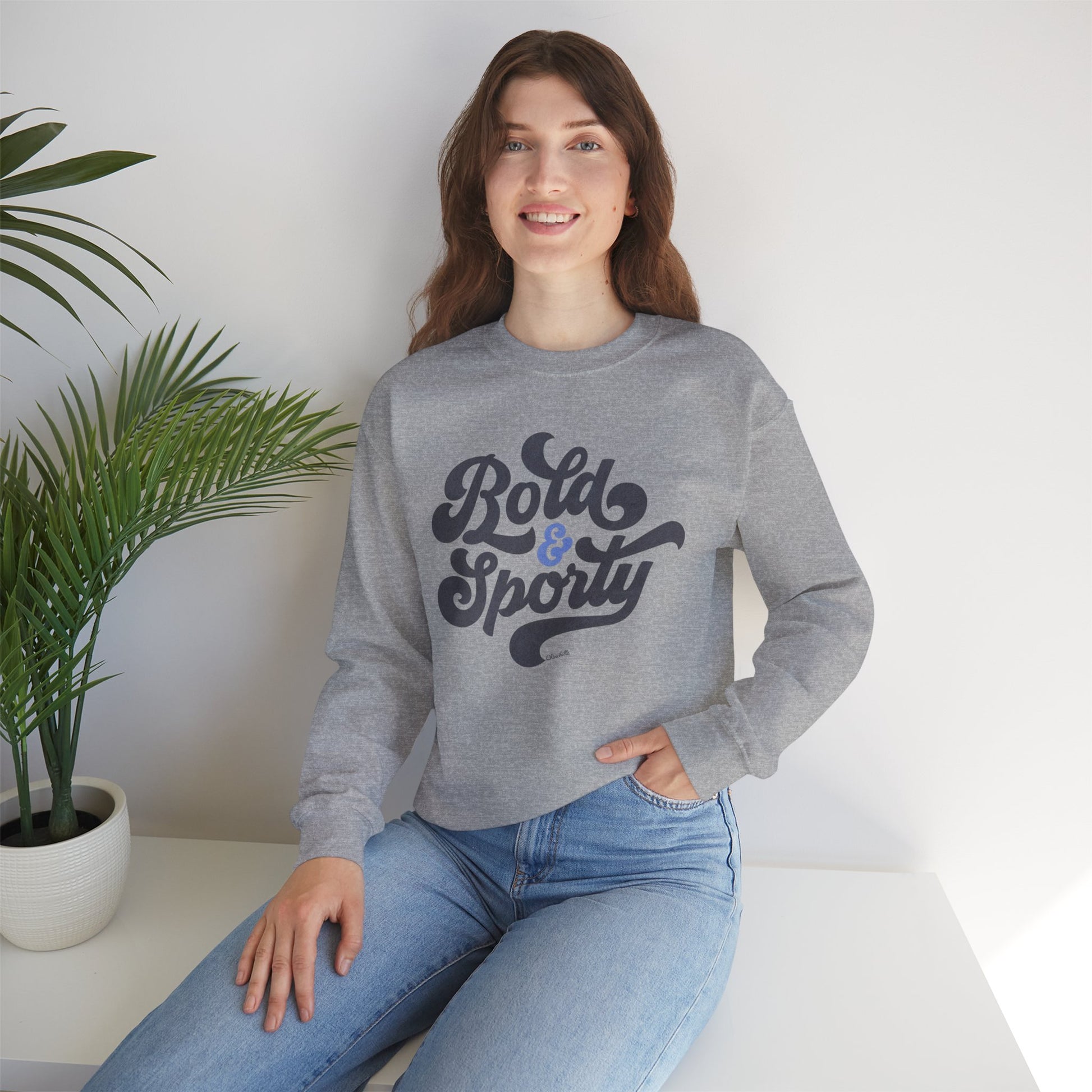 Woman wearing a gray sweatshirt with 'Bold & Sporty' text, sitting on a white surface with a plant in the background.