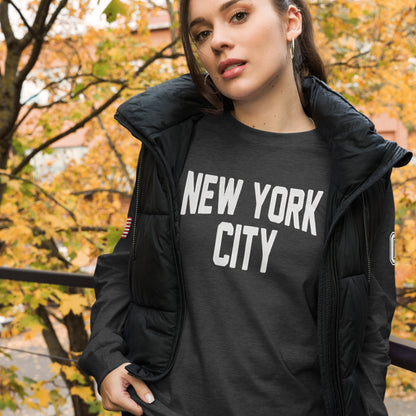 Woman wearing a black 'New York City' t-shirt with a black puffer vest outdoors with autumn trees.